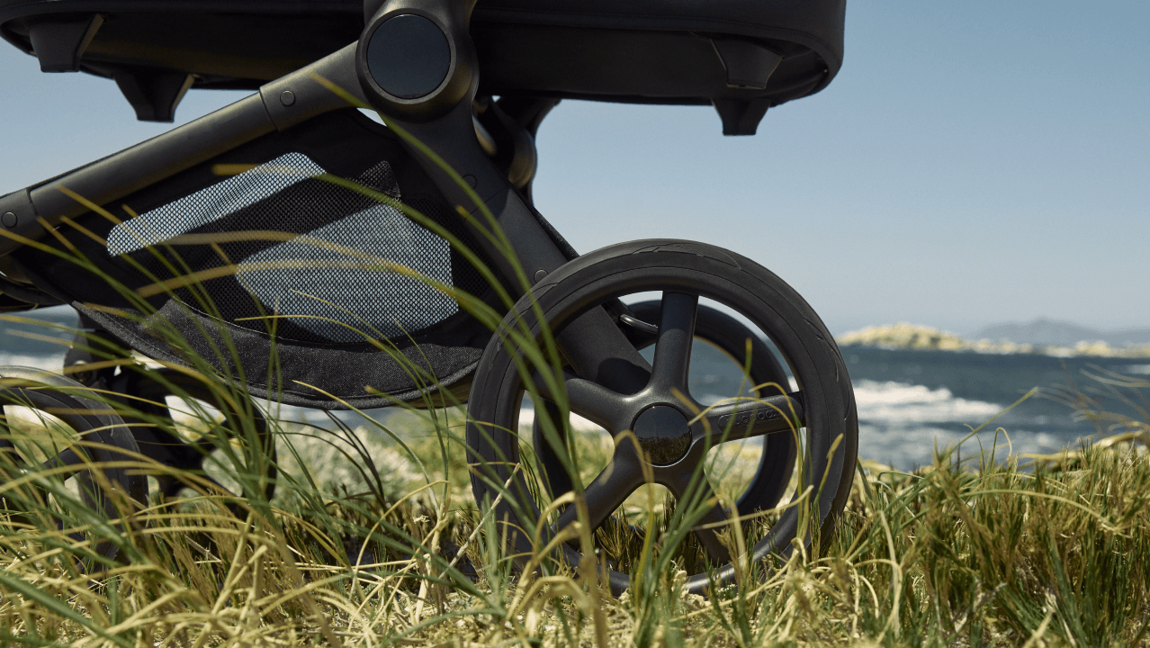Close-up of the all-terrain wheels of a Bugaboo stroller as it moves across a grassy area, with a scenic coastline and sea in the background.
