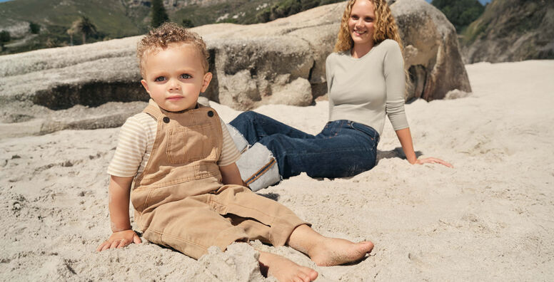 A baby sits barefoot in the sand with his mom behind him, on the beach near large rocks on a sunny day.