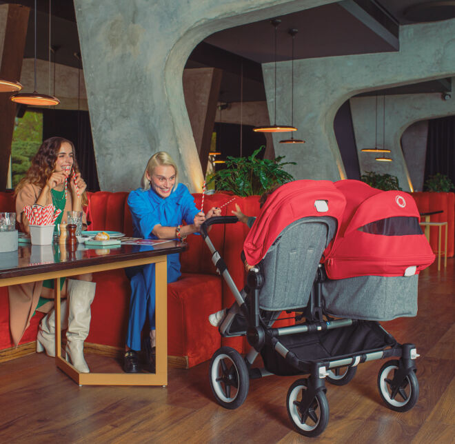 Una pareja se sienta en un restaurante y mira a sus bebés en un cochecito doble con un toldo rojo