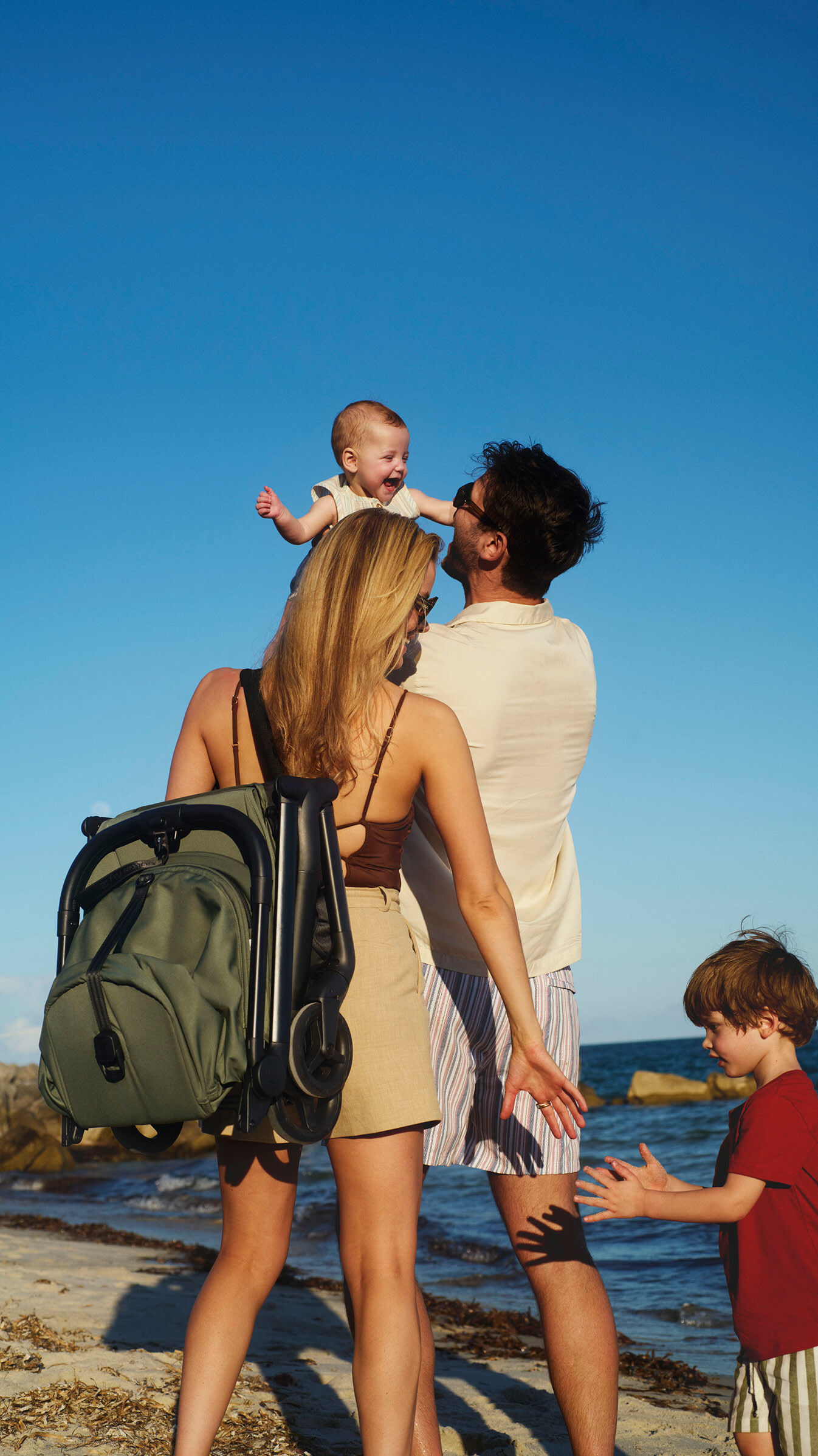 A happy family by the seaside. Mom takes the hand of their toddler while dad lifts their baby into the air. Mom carries a Bugaboo Butterfly 2 stroller on her shoulder with the carry strap.
