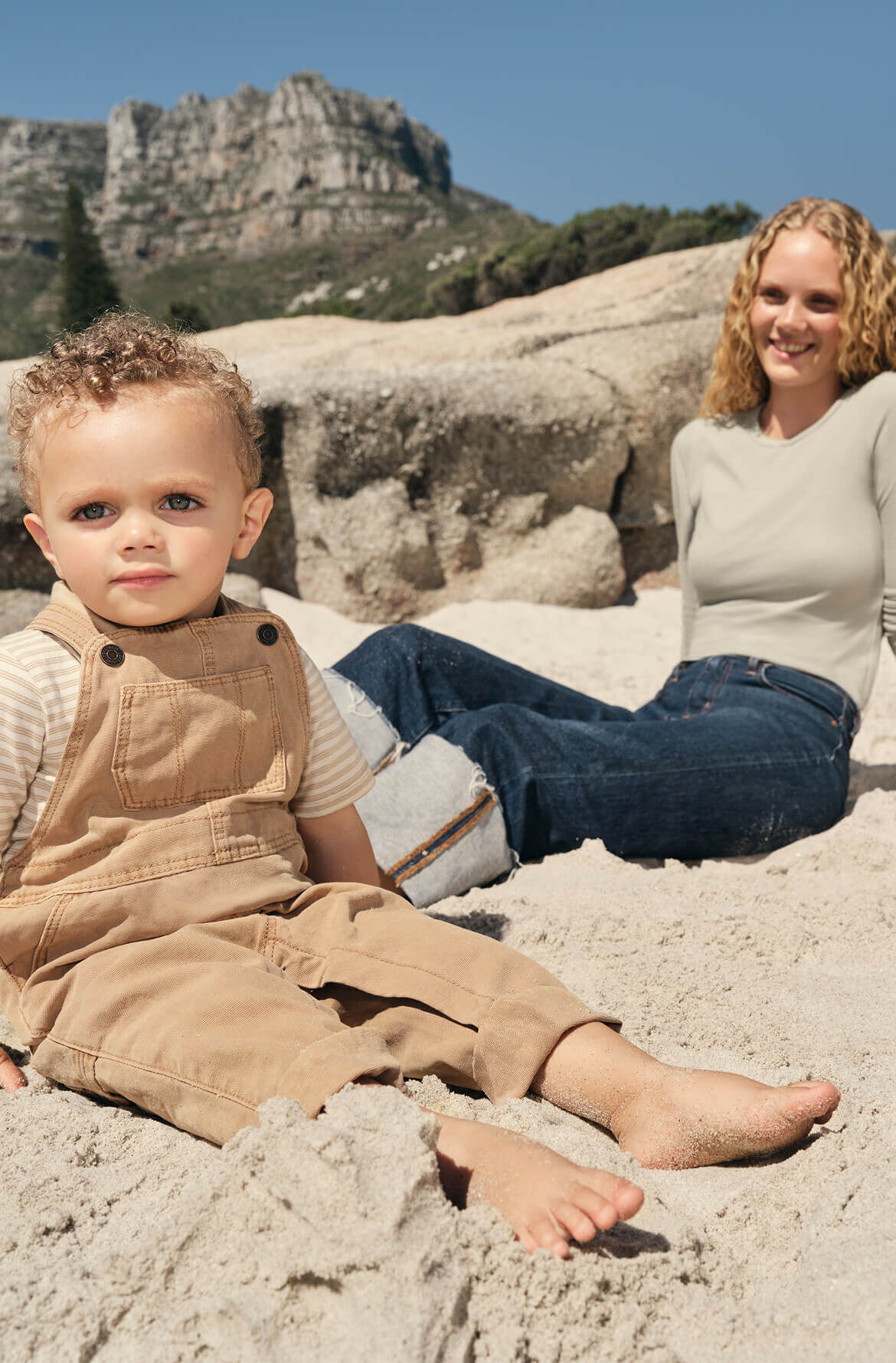 A baby sits barefoot in the sand with his mom behind him, on the beach near large rocks on a sunny day.