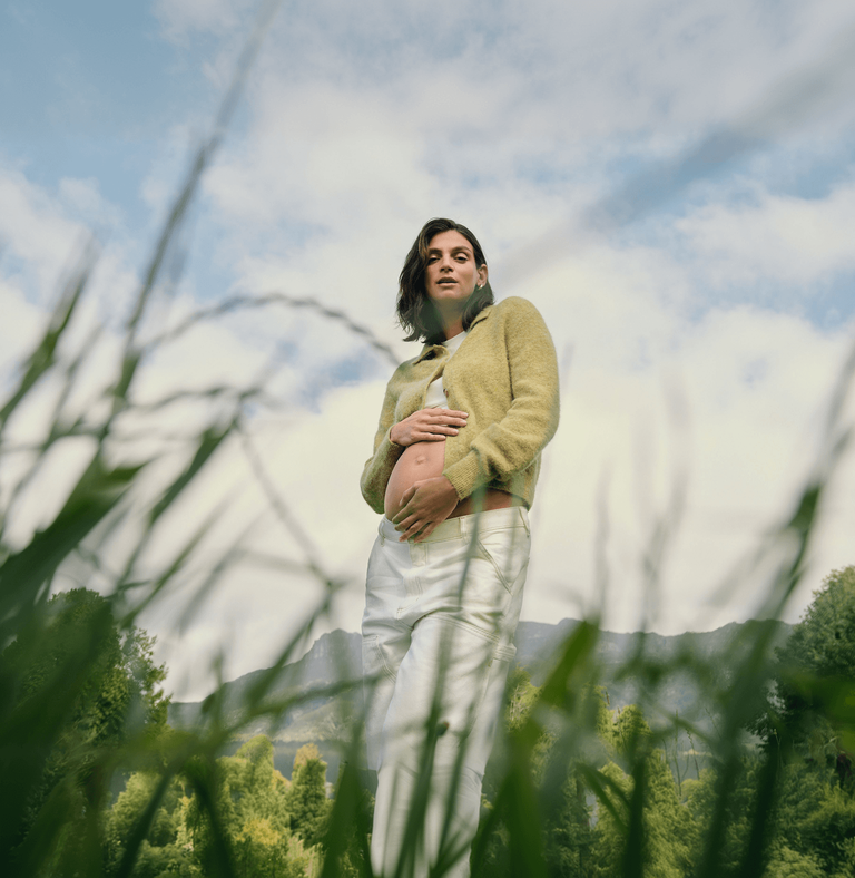 A pregnant mom stands in a grassy field with one hand on her bare baby bump, with mountains in the background and tall blades of grass in the foreground.