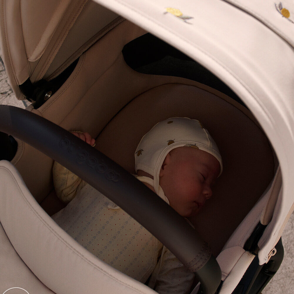 A baby sleeps inside a Bugaboo stroller's bassinet, shaded by the Bugaboo X Konges Sløjd Limited Edition sun canopy which features Konges Sløjd's signature lemon print.