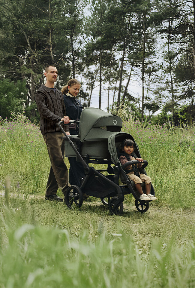 A happy family enjoys their day out. Mom and dad walk side by side, pushing a Bugaboo Kangaroo tandem stroller. One child sits in the seat, while another is hidden in the bassinet. The scene is surrounded by tall grass.