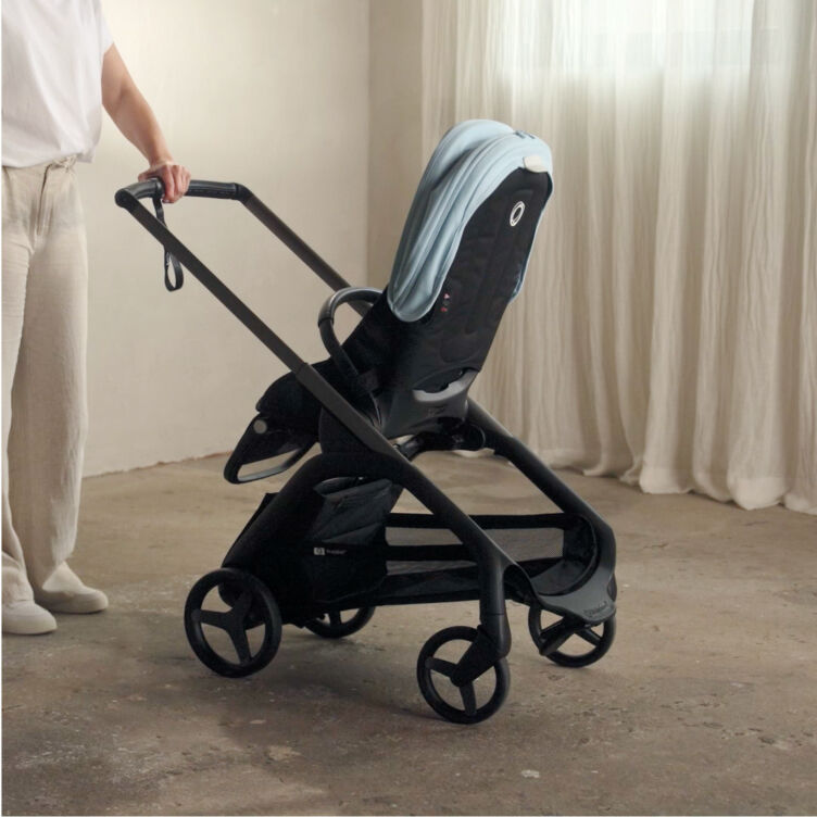 A Bugaboo expert stands next to a Bugaboo Dragonfly seat stroller with a light blue sun canopy, inside a softly lit room with white curtains.