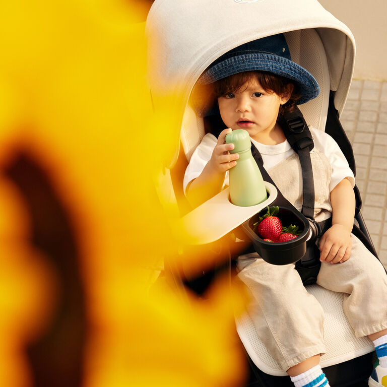 A baby wearing a denim hat and beige overalls sits in a Bugaboo stroller, in front of him is a snack tray with a water bottle and a compartment full of strawberries.