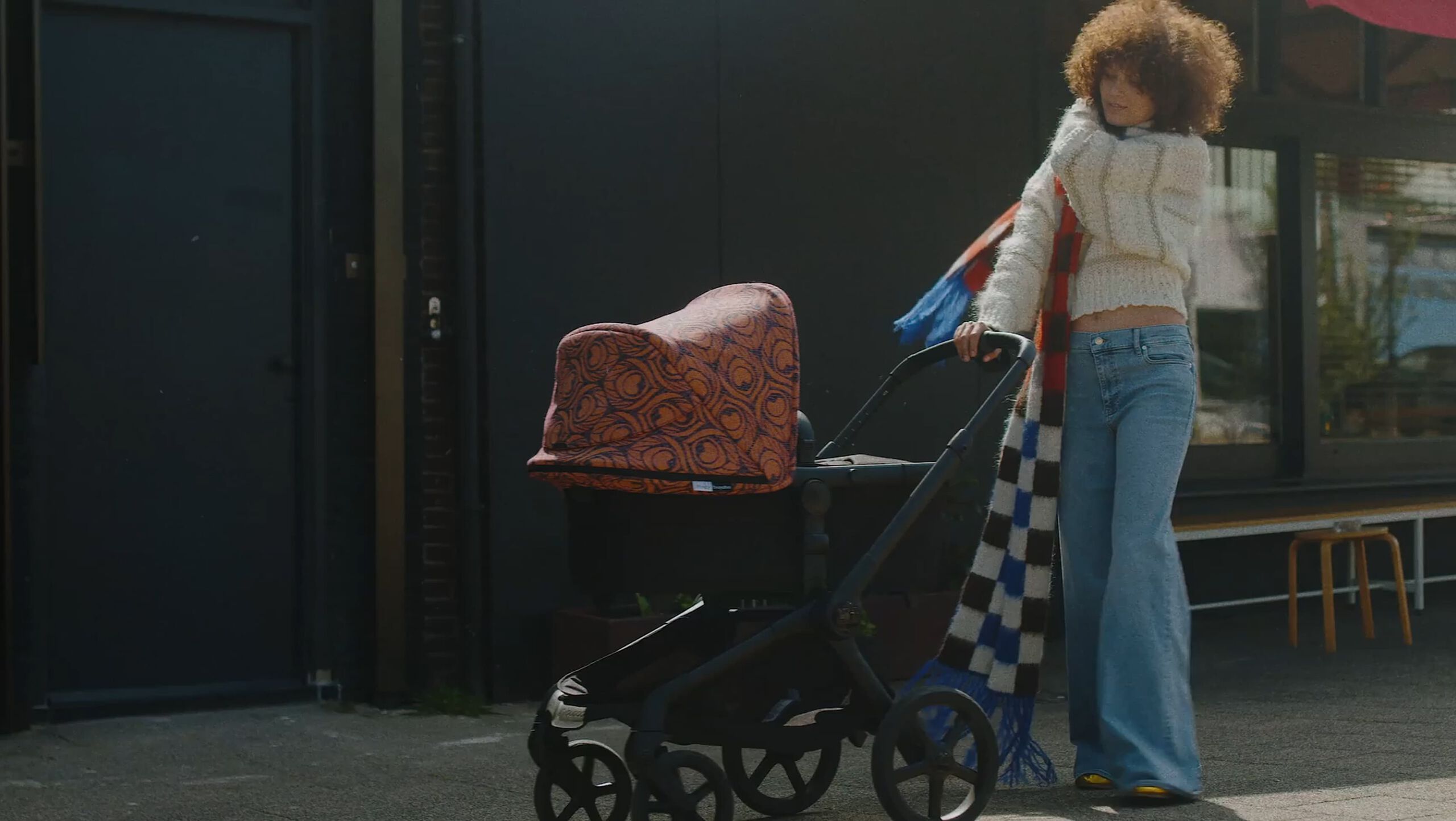 A stylish mom wearing a long checkered scarf strolls the street with her baby inside a Bugaboo Fox 5 Renew bassinet stroller. The stroller features Bugaboo X Artipoppe Limited Edition sun canopy with an exclusive patterned design.