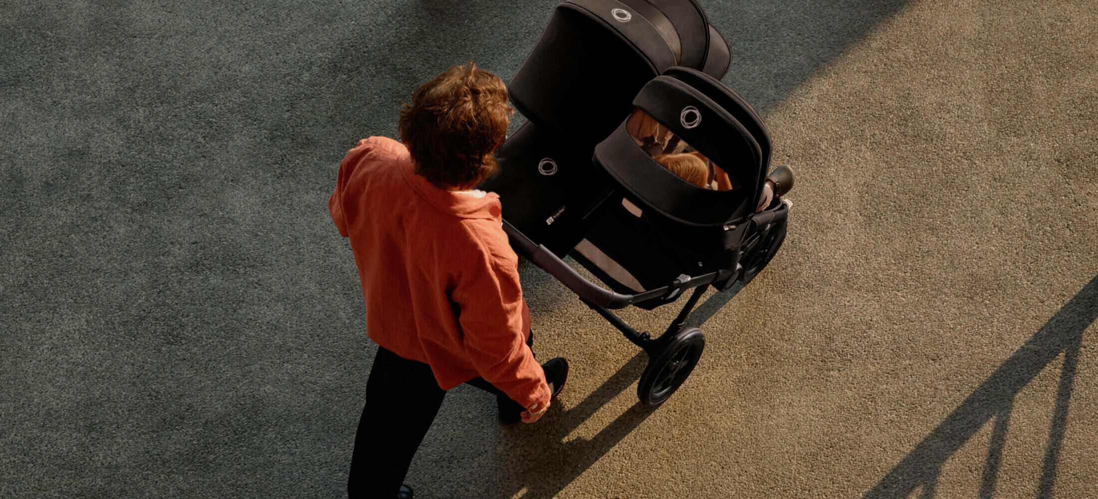 A top-down view of a father strolling with his two children in a Bugaboo Donkey 5 double stroller, with a seat and a bassinet in black fabrics. A toddler is visible through the peek-a-boo panel of the seat's sun canopy.