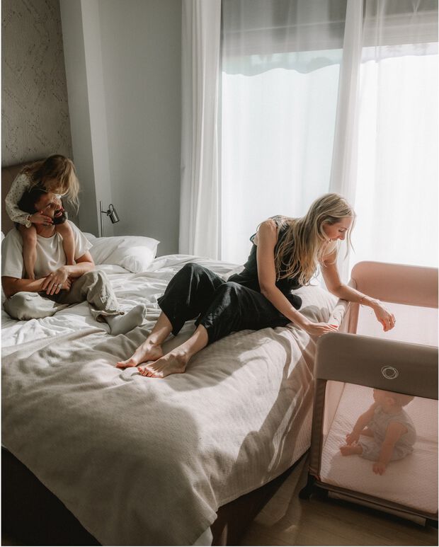 A dad, a mom, and an older child sit and play on the bed. The mom leans over to check on her baby in a Bugaboo Stardust travel cot, which has Desert Taupe fabrics.