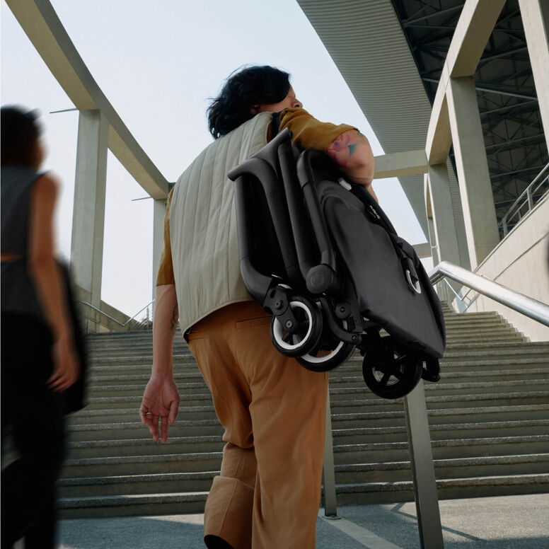 A dad carries a folded Bugaboo compact stroller on one shoulder as he heads towards the stairs.