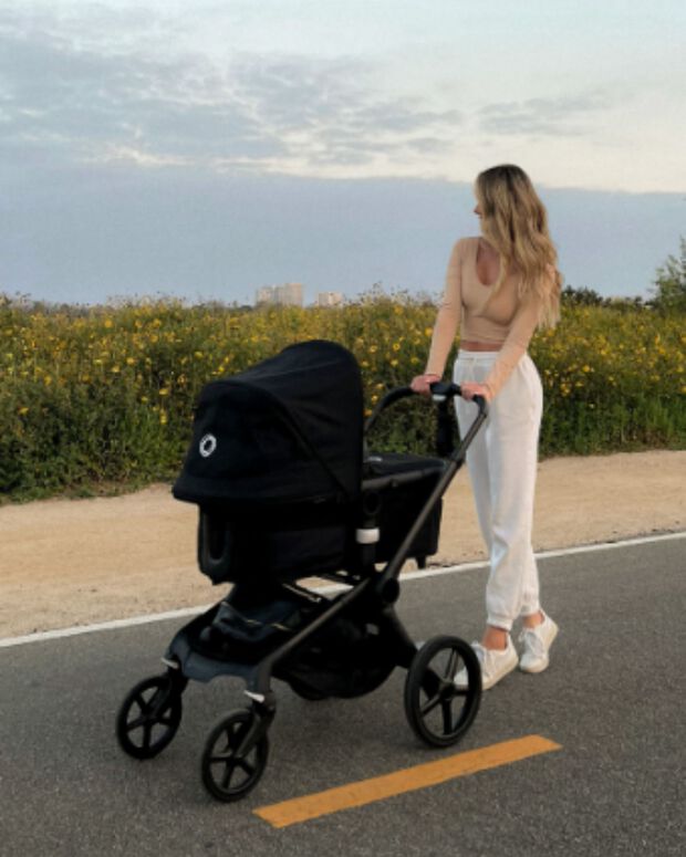 Une maman promène son bébé dans une Bugaboo Fox 5 sur une route pavée. En arrière-plan se trouvent un champ de fleurs et un ciel partiellement nuageux.