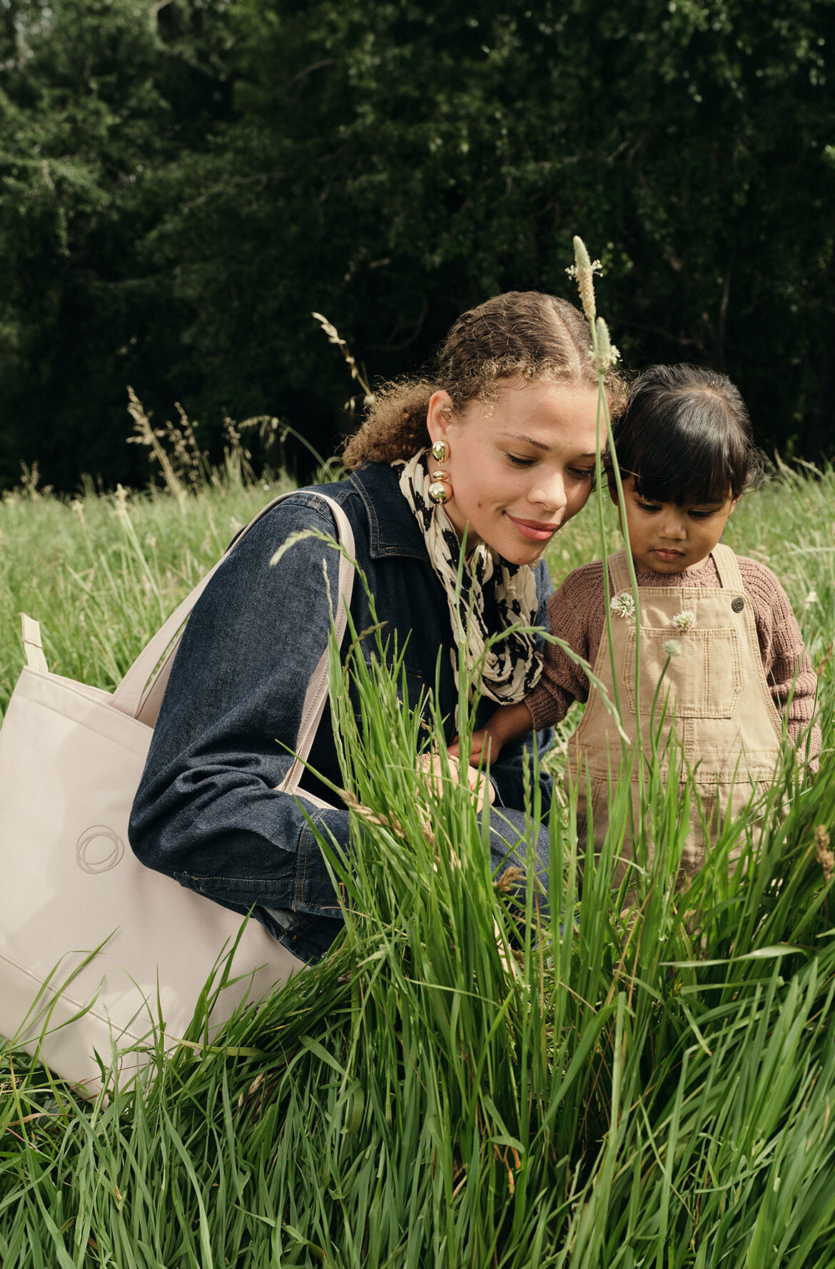 A mom and child crouch among tall grasses, curiously observing the plants. The mother carries a Bugaboo changing bag in Desert Taupe on her shoulder.