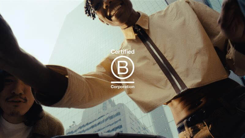 Point of view from a stroller looking up. A dad is looking down at his baby, smiling in happiness. Behind him are tall glass buildings. The B Corp logo sits in the middle of the picture.