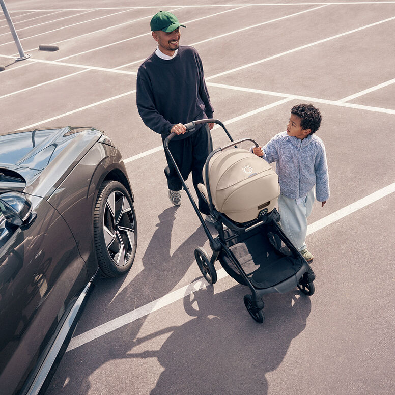 A dad pushes a Bugaboo stroller with a Bugaboo Otter by Nuna car seat attached to it across a car park, while his son walks alongside the stroller.