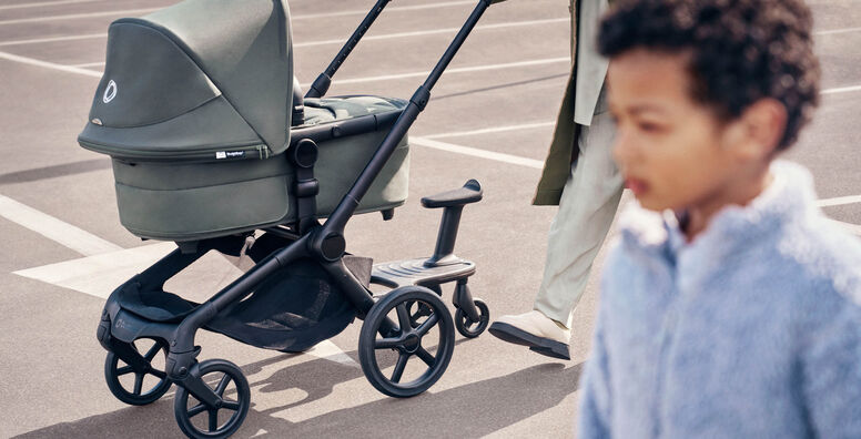 A parent pushes a Bugaboo Fox 5 Renew bassinet stroller with a Bugaboo wheeled board attached, while a child walks alongside.