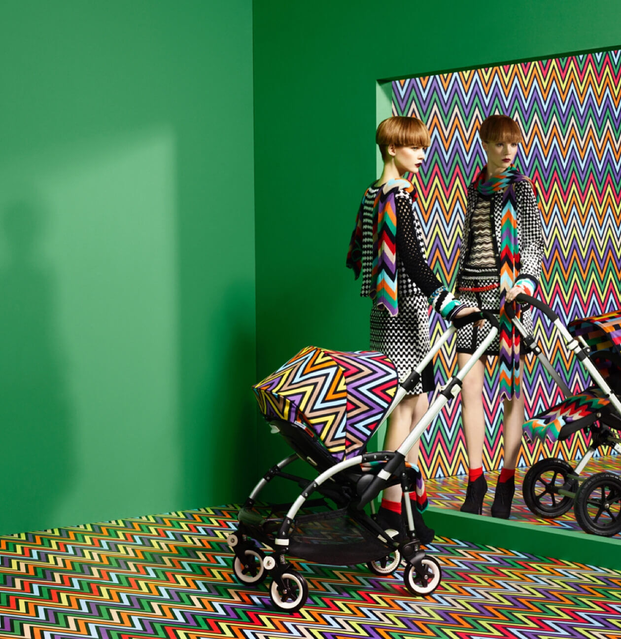 A female model stands next to a Bugaboo X Missoni Limited Edition stroller, looking into a mirror in a room with green walls. The stroller's sun canopy, the room's floor, and the wall reflected in the mirror are covered in Missoni's signature zigzag pattern.