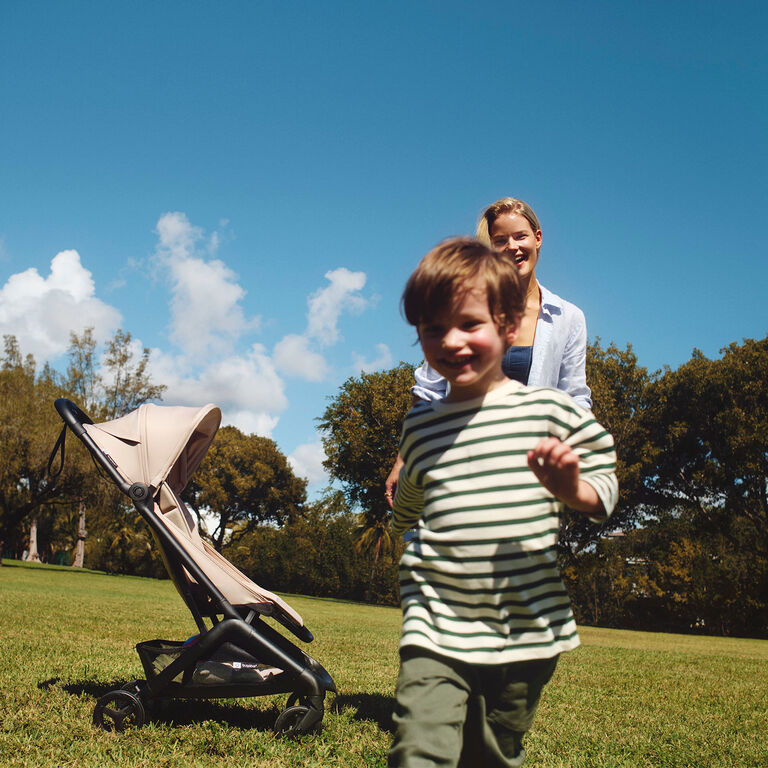 A boy runs on a grassy field. His smiling mom stands behind him, with a Bugaboo Butterfly 2 stroller in Desert Taupe fabric parked beside her.