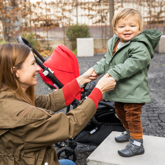 Mom and child holding hands while the child stands on a concrete block; behind them is the Bugaboo Bee 6 with red sun canopy.