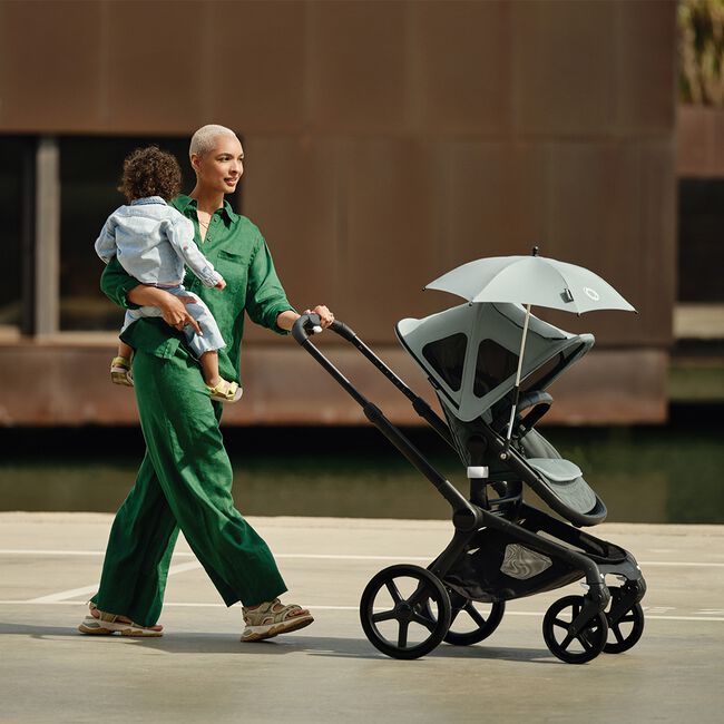 Mamma che tiene in braccio un bambino mentre spinge con l'altro braccio un passeggino Bugaboo Fox 5 dotato di diversi accessori.