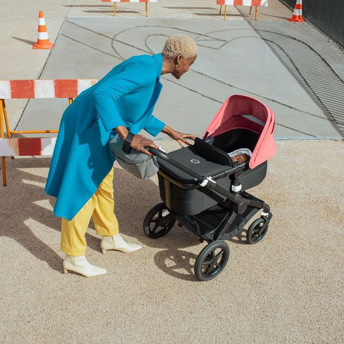 A woman wearing yellow trousers and a blue coat leaning over to check on a baby lying in the Bugaboo Fox carrycot and pushchair seat
